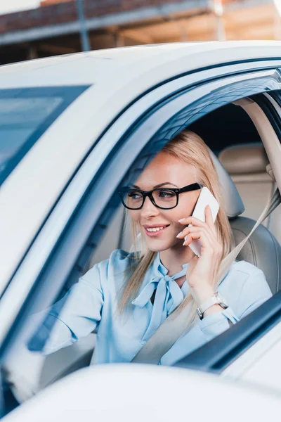 Blonde businesswoman in eyeglasses talking on smartphone while driving car — Stock Photo