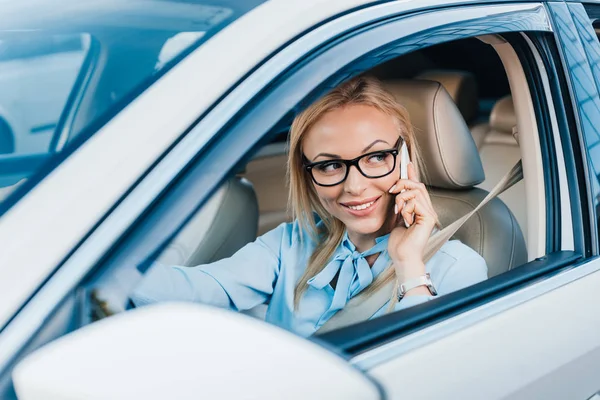 Smiling businesswoman in eyeglasses talking on smartphone while driving car — Stock Photo