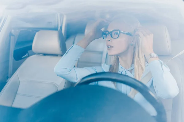 Blonde businesswoman in eyeglasses looking at rear view mirror in car — Stock Photo