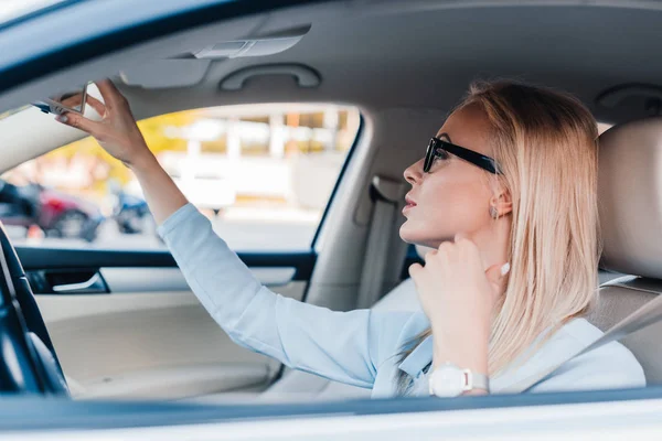 Vista laterale della donna d'affari bionda negli occhiali guardando lo specchio retrovisore in auto — Foto stock