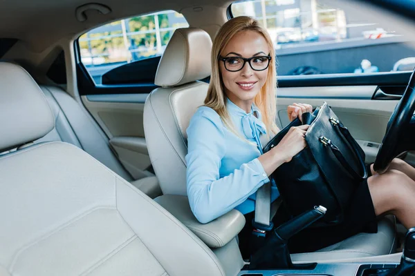 Smiling blonde businesswoman in eyeglasses checking bag in car — Stock Photo