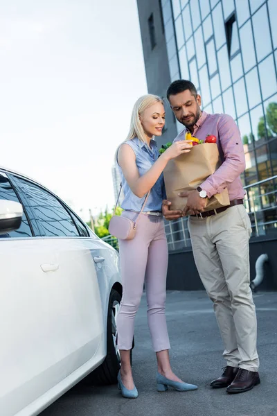 Married couple with paper bag full of healthy food on parking near car — Stock Photo