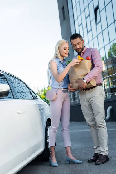 Casal com saco de papel cheio de comida saudável no estacionamento perto do carro — Fotografia de Stock