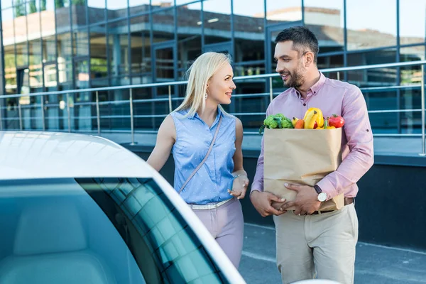 Portrait of married couple with paper bag full of healthy food on parking near car — Stock Photo