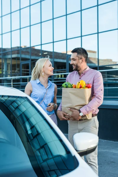 Portrait of married couple with paper bag full of healthy food on parking near car — Stock Photo