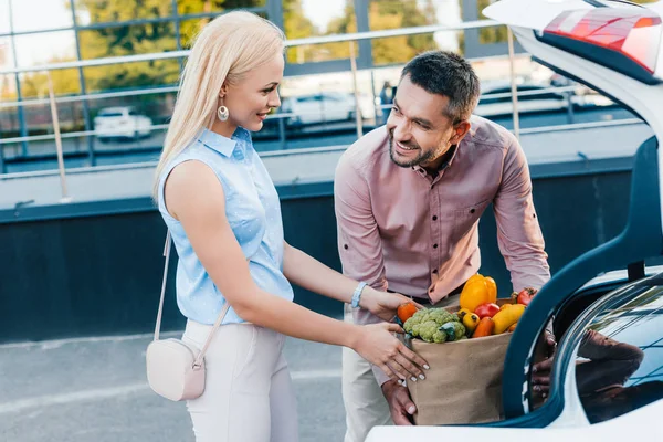 Portrait of married couple putting paper bag full of healthy food into car on parking — Stock Photo