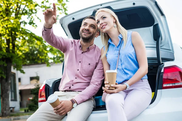 Portrait of smiling couple with coffee to go sitting on car and looking away — Stock Photo