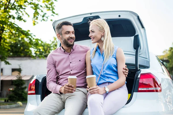 Portrait de couple souriant avec café pour aller s'asseoir sur la voiture et se regarder — Photo de stock