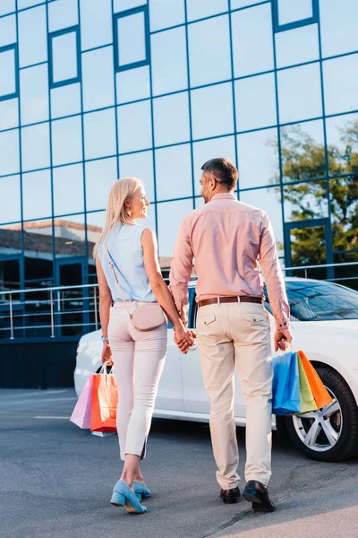 Vista trasera de la pareja casada con bolsas de compras cogidas de la mano y caminando al coche en la calle - foto de stock