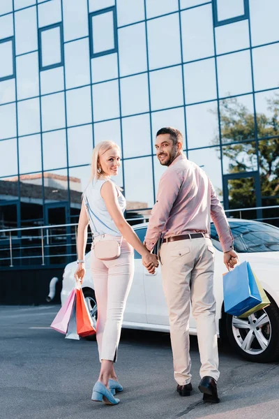 Visão traseira do casal com sacos de compras de mãos dadas e caminhando para o carro na rua — Fotografia de Stock