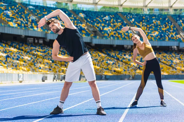 Joven pareja deportiva haciendo curva lateral antes de entrenar en pista de atletismo en el estadio deportivo - foto de stock