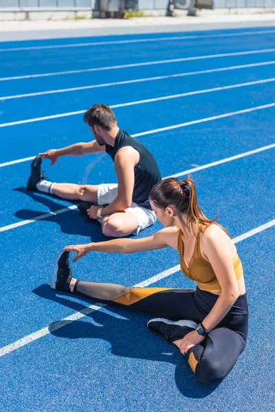 Visão de alto ângulo de jovens atléticos joggers masculinos e femininos sentados em pista de corrida e alongamento — Fotografia de Stock