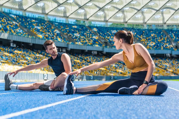 Joggers masculinos y femeninos en forma joven sentados en pista de atletismo y estirándose en el estadio deportivo - foto de stock