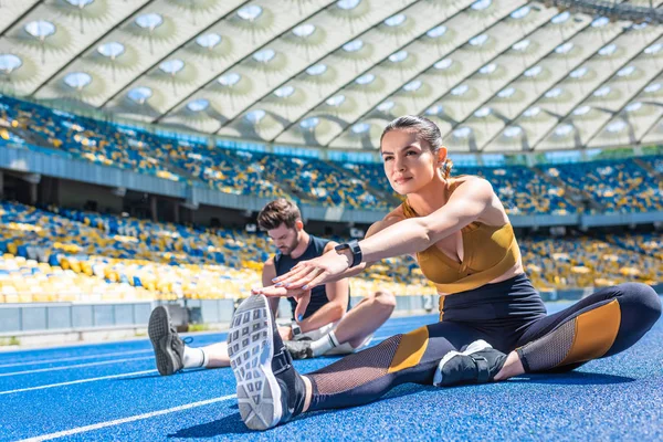 Jóvenes corredores masculinos y femeninos sentados en pista de atletismo y estirándose en el estadio deportivo - foto de stock