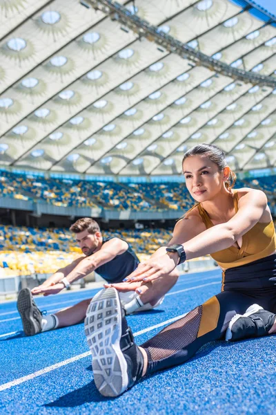 Young attractive couple sitting on running track and stretching at sports stadium — Stock Photo