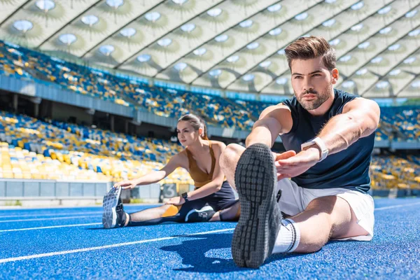 Young fit couple sitting on running track and stretching at sports stadium — Stock Photo