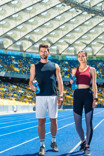 Jovem casal esportivo com garrafas de água em pé na pista de corrida no estádio de esportes — Fotografia de Stock