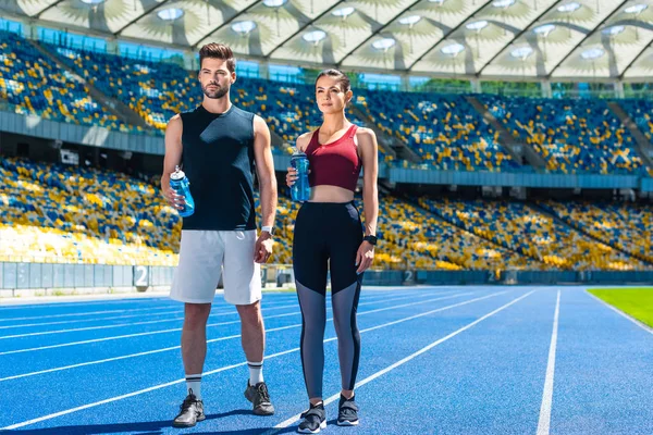 Молодая спортивная пара с бутылками воды отдыхает на беговой дорожке на спортивном стадионе — стоковое фото