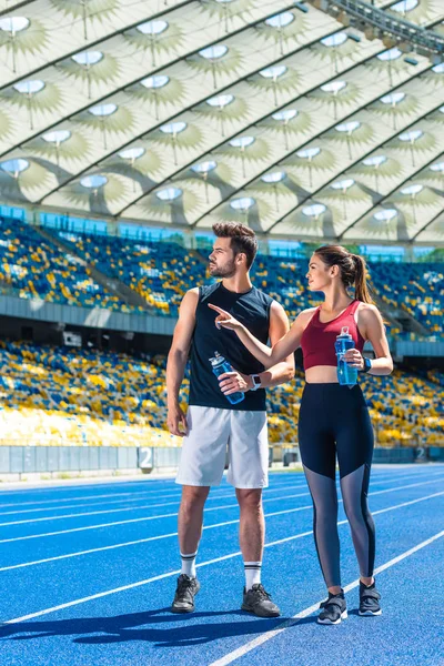 Pareja de jóvenes en forma con botellas de agua charlando en pista de atletismo en el estadio deportivo - foto de stock