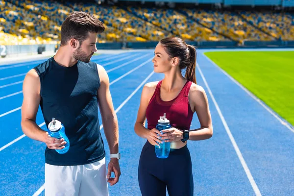 Joven pareja deportiva con botellas de agua charlando en pista de atletismo en el estadio deportivo - foto de stock