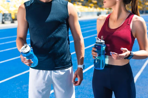 Tiro recortado de pareja en forma con botellas de agua en pista de atletismo en el estadio deportivo - foto de stock