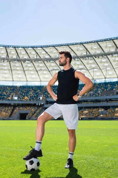 Серьезный молодой футболист, стоящий на спортивном стадионе с мячом и смотрящий в сторону — стоковое фото