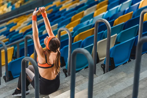 Vista de ángulo alto de la hermosa mujer joven estirándose mientras está sentado en las escaleras en el estadio de deportes - foto de stock