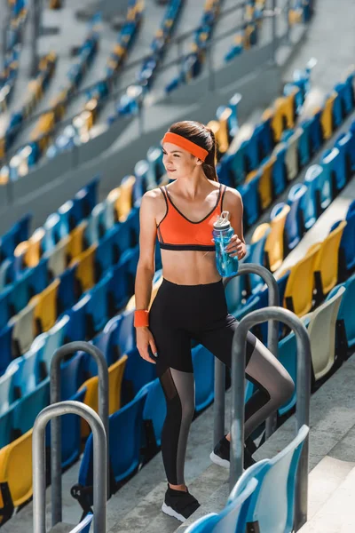 Высокий угол обзора счастливой молодой женщины с бутылкой воды, стоящей на трибунах на спортивном стадионе — стоковое фото