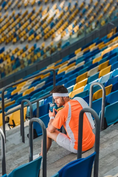 Cansado joven deportista sentado en las escaleras en el estadio de deportes con botella de agua - foto de stock