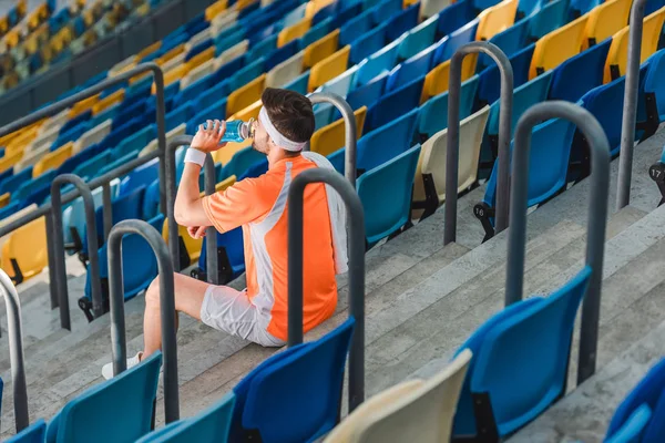 Cansado joven deportista beber agua mientras está sentado en las escaleras en el estadio de deportes - foto de stock