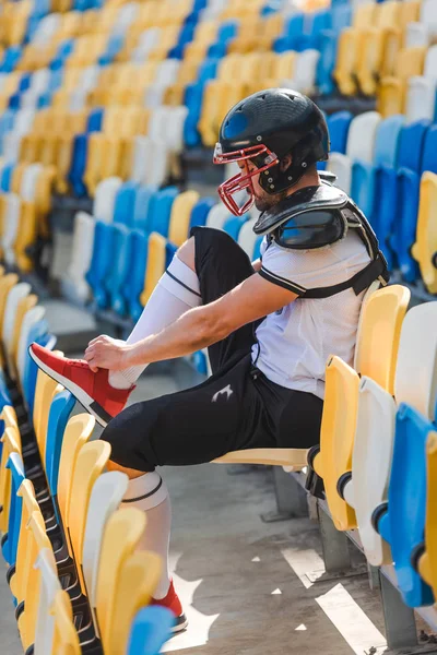 Vista lateral del joven futbolista americano sentado en tribunas en el estadio deportivo y atando zapatos - foto de stock