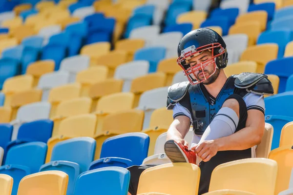 Guapo joven jugador de fútbol americano sentado en tribunas en el estadio de deportes y zapatos de cordones - foto de stock