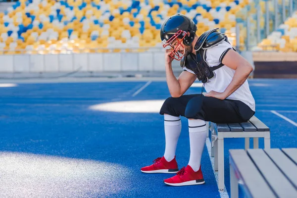 Vista lateral del joven jugador de fútbol americano sosteniendo la jaula del casco mientras está sentado en el estadio deportivo - foto de stock