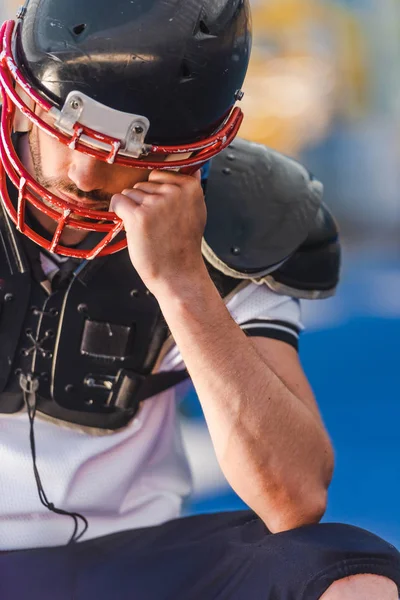 Junger amerikanischer Fußballspieler hält Käfig mit Helm während er im Sportstadion sitzt — Stockfoto