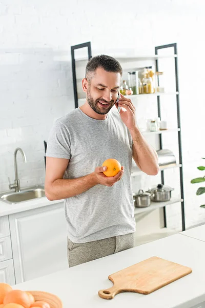 Улыбающийся мужчина разговаривает на смартфоне и держит апельсин на кухне — стоковое фото