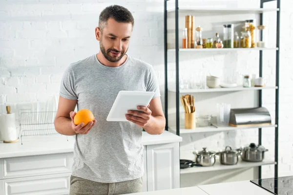 Adult man with digital tablet holding orange and looking for recipe on kitchen — Stock Photo