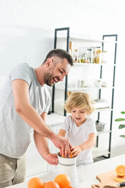 Feliz padre e hijo pequeño haciendo jugo de naranja fresco por exprimidor en la mesa en la cocina - foto de stock