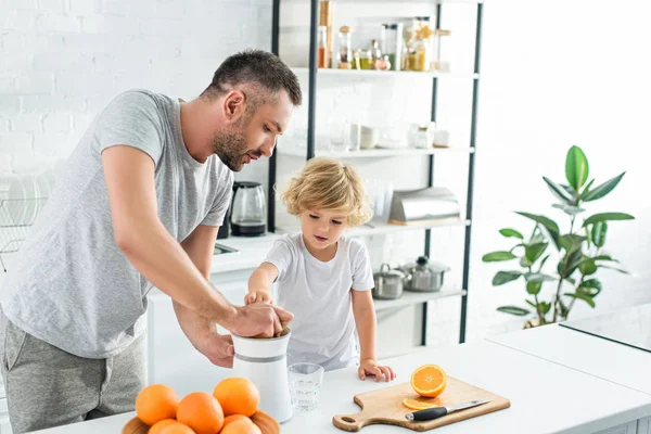 Vater und kleiner Sohn bereiten frischen Orangensaft per Pressmaschine auf Tisch bei Sohn in der Küche zu — Stockfoto