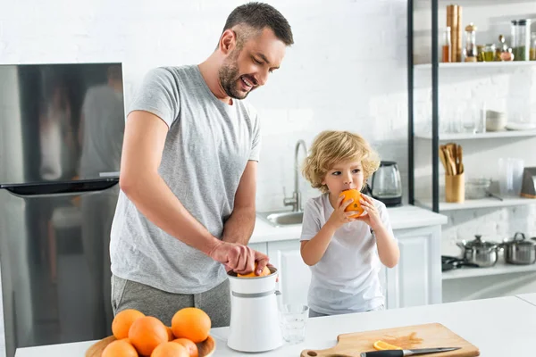Padre haciendo jugo de naranja por exprimidor mientras su pequeño hijo comiendo naranja en la cocina - foto de stock