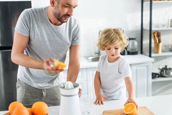 Pequeño niño dando padre naranja para hacer jugo fresco en la cocina - foto de stock