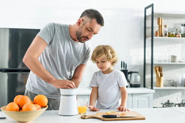 Pai focado e filho pequeno fazendo suco de laranja fresco pelo espremedor na mesa na cozinha — Fotografia de Stock