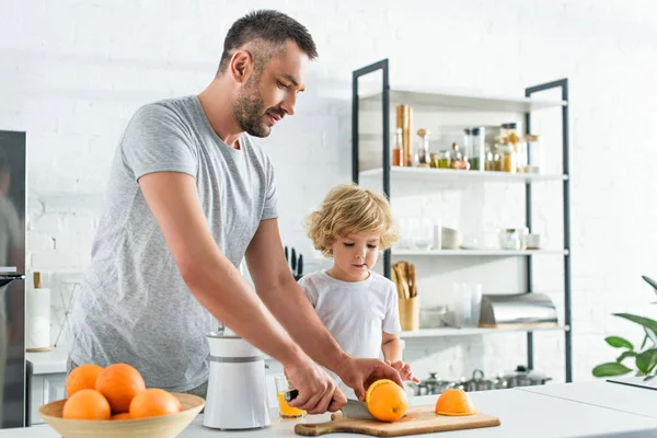 Homem cortando laranja por faca para fazer suco fresco, enquanto seu filho de pé perto na cozinha — Fotografia de Stock