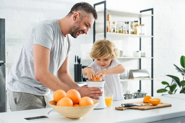 Padre sorridente e piccolo figlio che fa succo d'arancia fresco da spremiagrumi sul tavolo in cucina — Foto stock