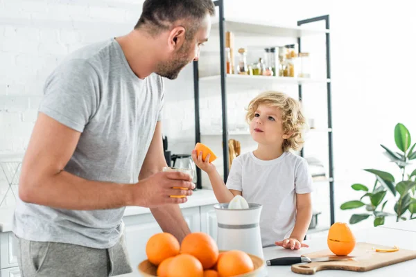 Hijo y padre mirándose el uno al otro después de hacer jugo de naranja fresco en kithen - foto de stock