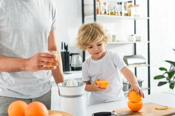 Imagen recortada del padre con un vaso de jugo fresco y su hijo tomando naranja de la tabla de cortar en la cocina - foto de stock