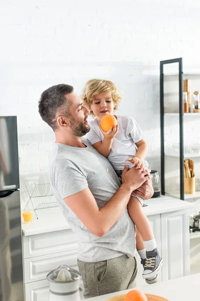 Hombre adulto sosteniendo pequeño hijo con naranja en las manos en la cocina - foto de stock
