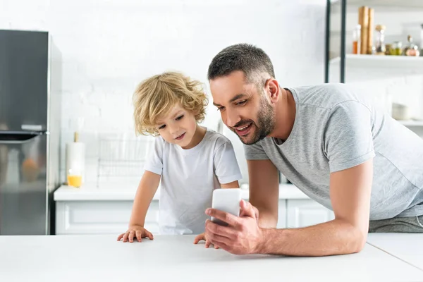Happy man showing smartphone screen to adorable son at kitchen — Stock Photo
