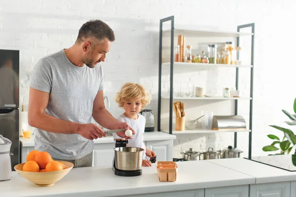 Vista lateral del hombre rompiendo huevo por cuchillo para hacer masa mientras su hijo de pie cerca de la cocina - foto de stock