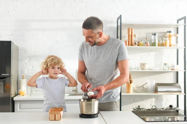 Menino fechando ouvidos por dedos, enquanto seu pai quebrando para fazer massa na cozinha — Fotografia de Stock