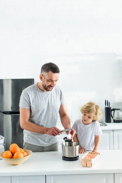 Человек с сыном разбивают яйцо ножом в миску на кухне — стоковое фото